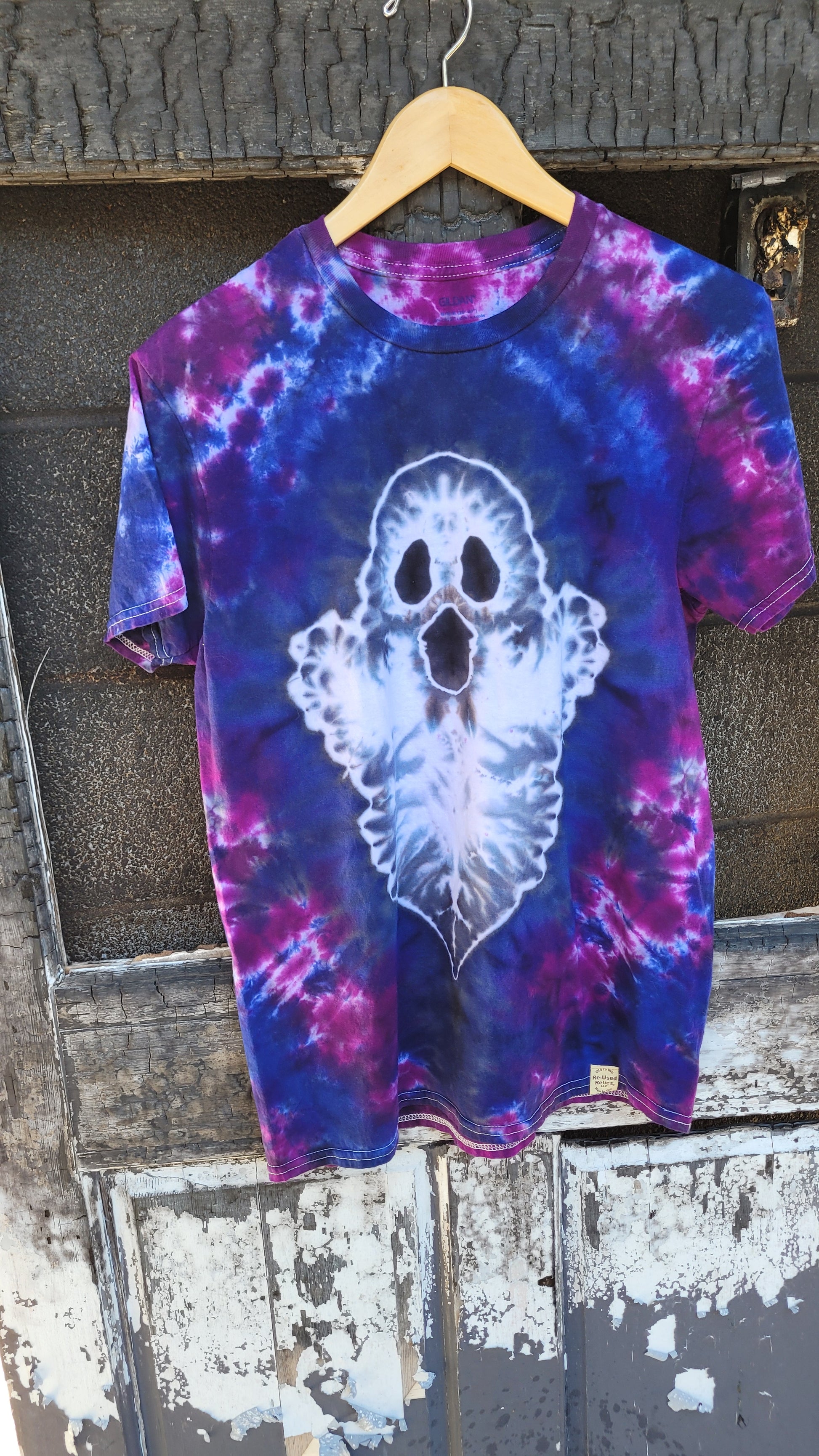 A short-sleeve crewneck t-shirt with a blue, purple, and pink tie-dye pattern, featuring a large white ghost graphic on the front. The shirt is hanging on a wooden hanger against a weathered, rustic wooden door.