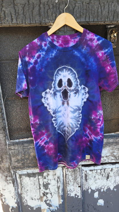 A short-sleeve crewneck t-shirt with a blue, purple, and pink tie-dye pattern, featuring a large white ghost graphic on the front. The shirt is hanging on a wooden hanger against a weathered, rustic wooden door.