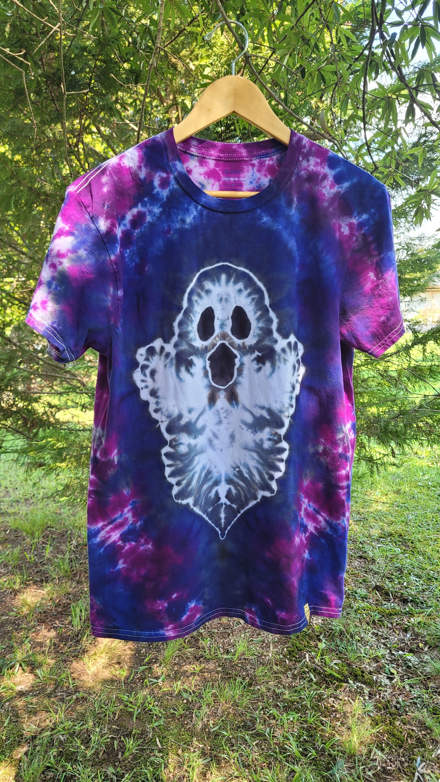A short-sleeve tie-dye t-shirt hanging on a wooden hanger outdoors. The shirt features a purple and blue spiral tie-dye pattern with a large white ghost graphic on the front.