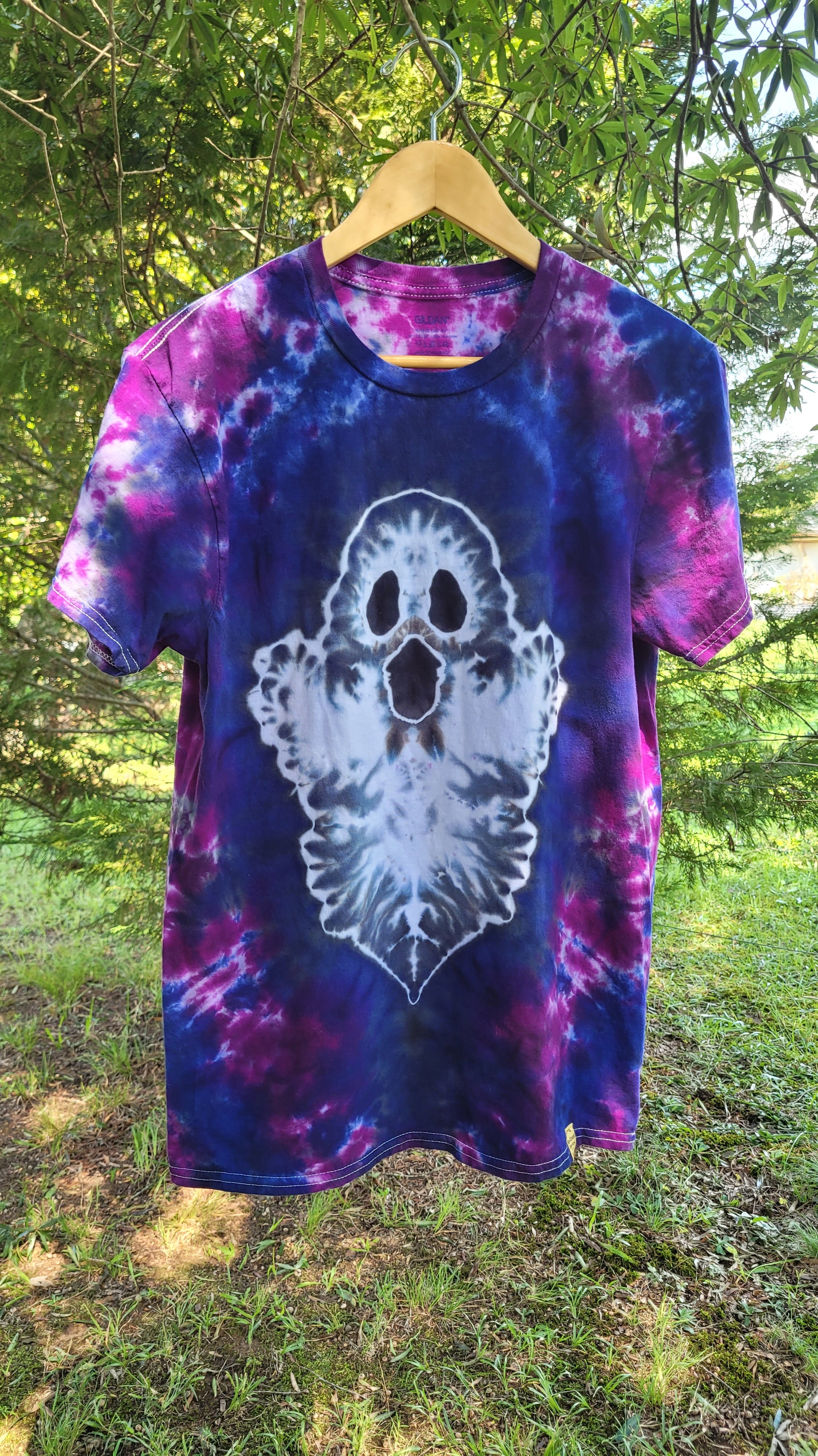 A short-sleeve tie-dye t-shirt hanging on a wooden hanger outdoors. The shirt features a purple and blue spiral tie-dye pattern with a large white ghost graphic on the front.
