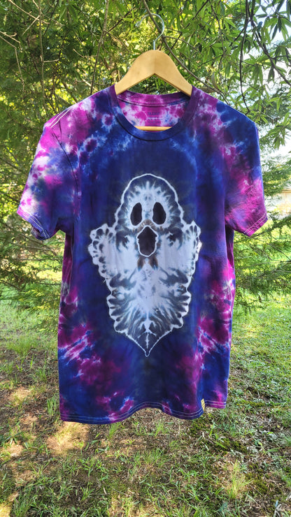 A short-sleeve tie-dye t-shirt hanging on a wooden hanger outdoors. The shirt features a purple and blue spiral tie-dye pattern with a large white ghost graphic on the front.